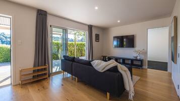 Ein modernes Wohnzimmer mit einem dunklen Sofa, Holzboden, einem an der Wand montierten Fernseher und einer Glastür zum Balkon.