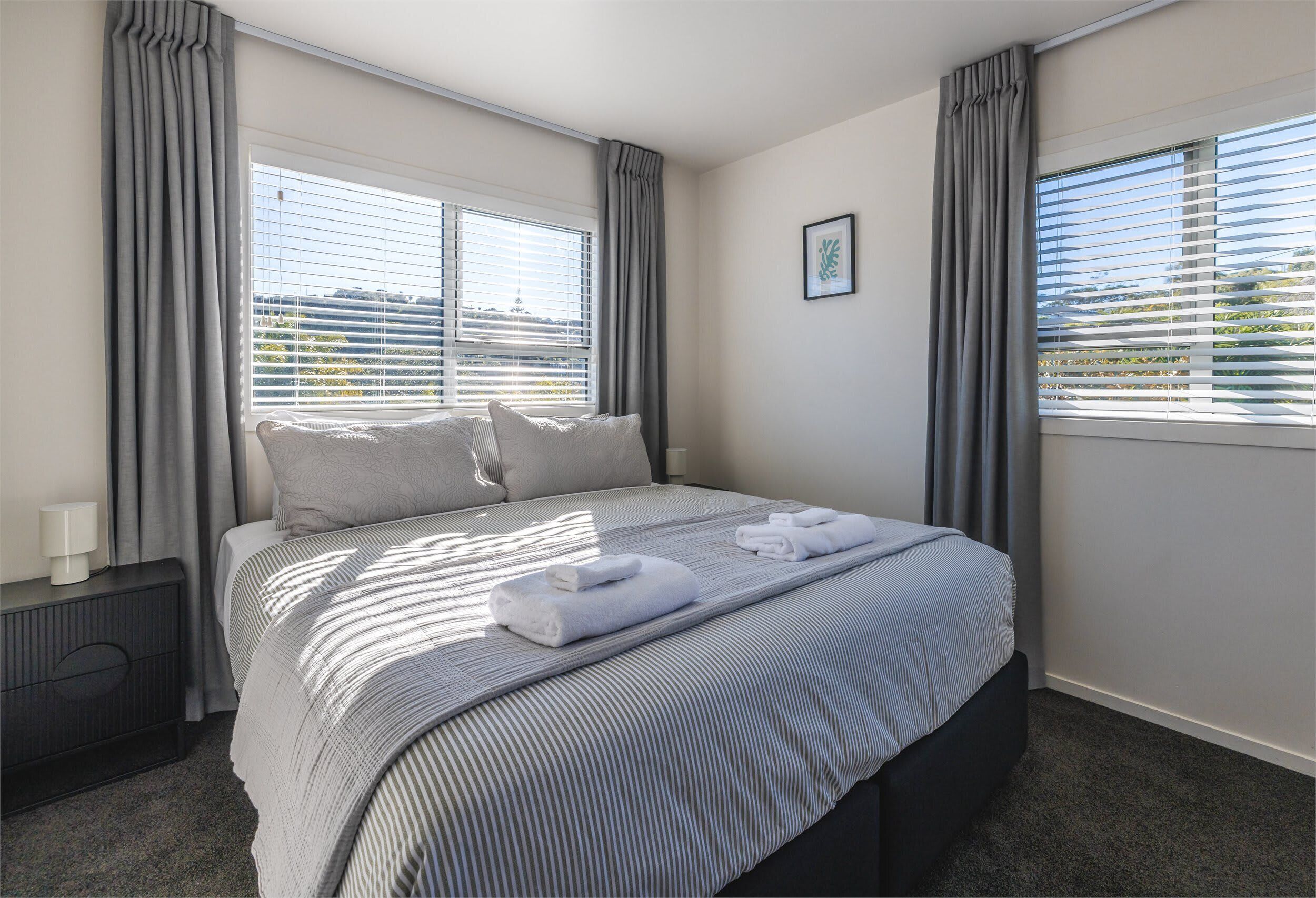 A hotel room with a bed, curtains, a window with blinds, a bedside table, and a picture on the wall.