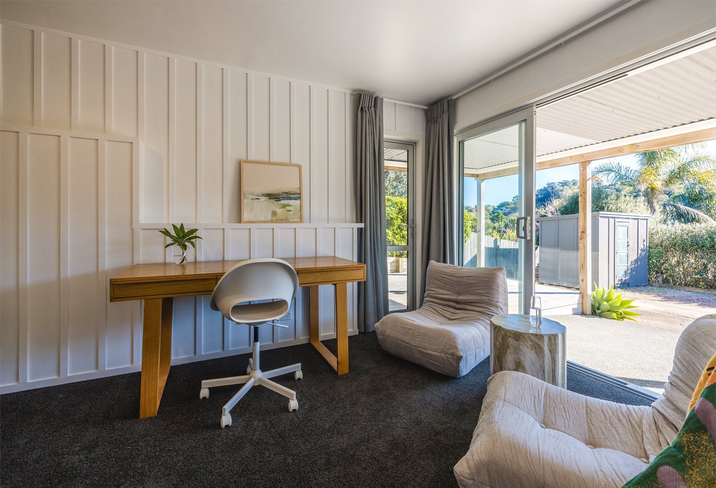 A modern room with a desk, chair, and a sofa. There is a window with curtains, a plant on the desk, and a view of the outdoors.