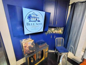 A room with a television mounted on the wall displaying 'The Blue Note House' sign. There is a small table with a record player and vinyl records, and a chair in the corner.