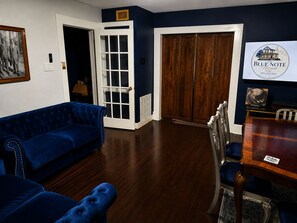 A room with a blue velvet sofa, a wooden dining table with chairs, a closed wooden door, and a framed picture on the wall. - The Blue Note House: Historic 6-bedroom Marvelous Music-Themed Gulfport Getaway (Gulfport)
