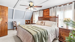 Ein Schlafzimmer mit einem Bett, einem Holzkopfende, einem Deckenventilator, einem Nachttisch, einer Lampe, einem Sessel und einem Fenster mit Vorhängen.