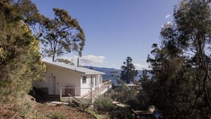 A two-story house with a balcony, surrounded by trees and a view of the sea. - Graceys Beach House Dennes Point (Kingborough)