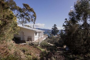 A two-story house with a balcony, surrounded by trees and a view of the sea. - Graceys Beach House Dennes Point (Kingborough)
