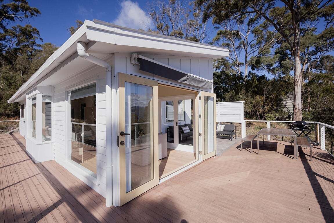Un bâtiment moderne, blanc, de plain-pied, doté de grandes portes et fenêtres en verre, d’une terrasse avec des meubles d’extérieur et offrant une vue dégagée sur les arbres environnants.