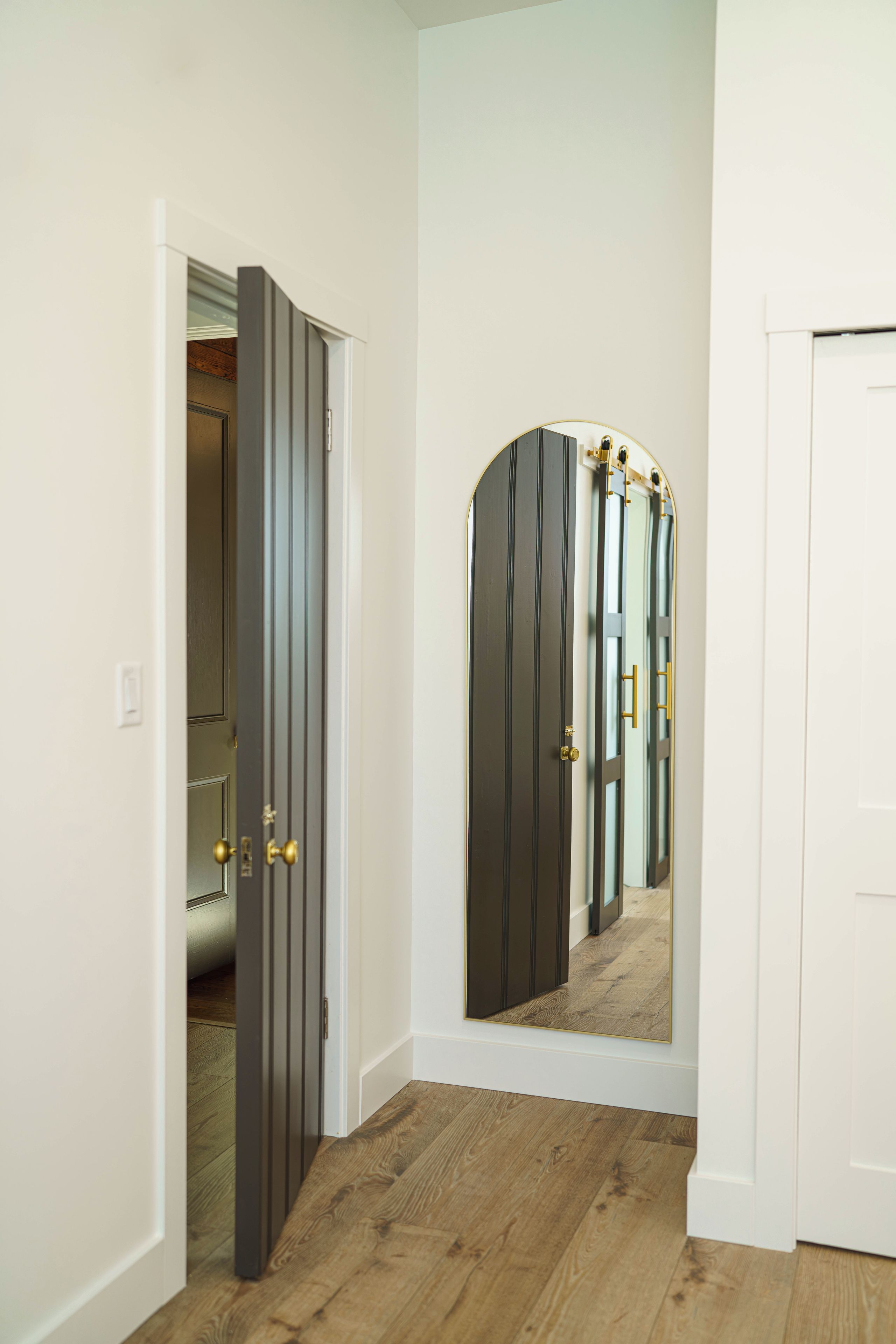 A hallway with a wooden floor, a white door, and a mirror reflecting a door with a golden handle.