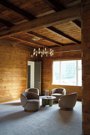 Una habitación con paredes y techo de madera, un espejo grande, dos sillones, una mesita redonda y una ventana con vistas a edificios.