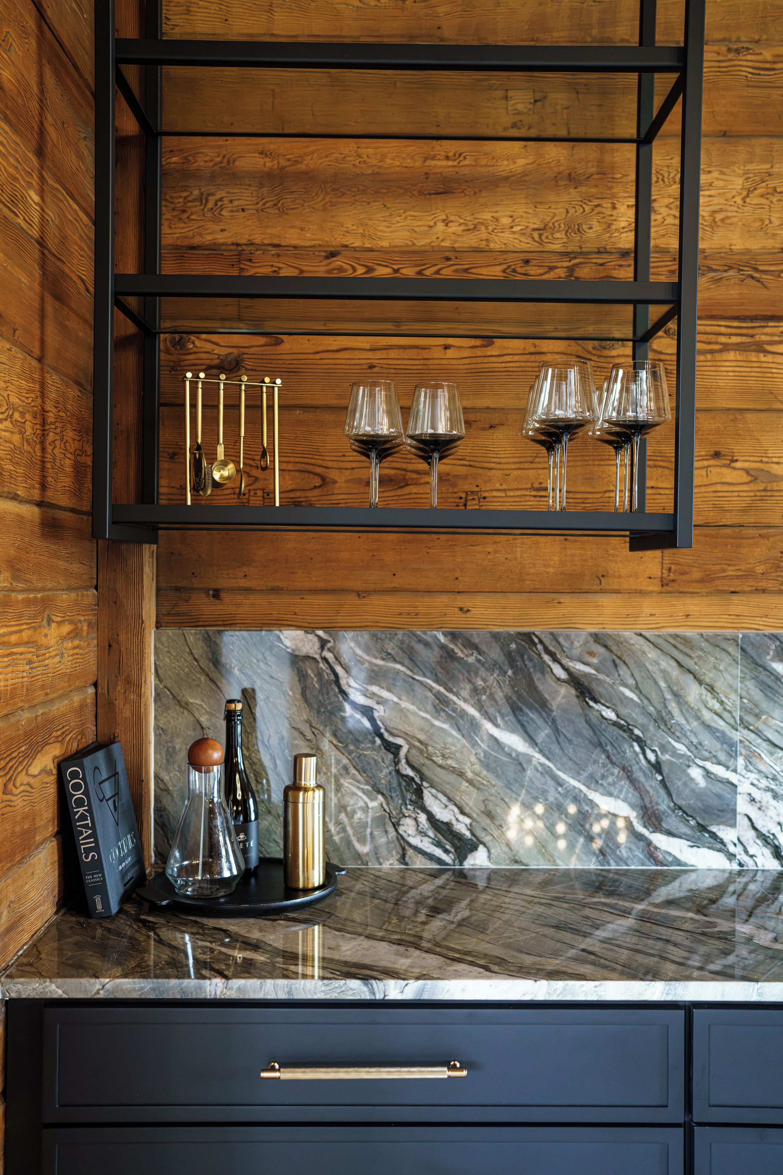 Una cocina con estante de madera que sostiene copas de vino y encimera de mármol con una botella y un libro.