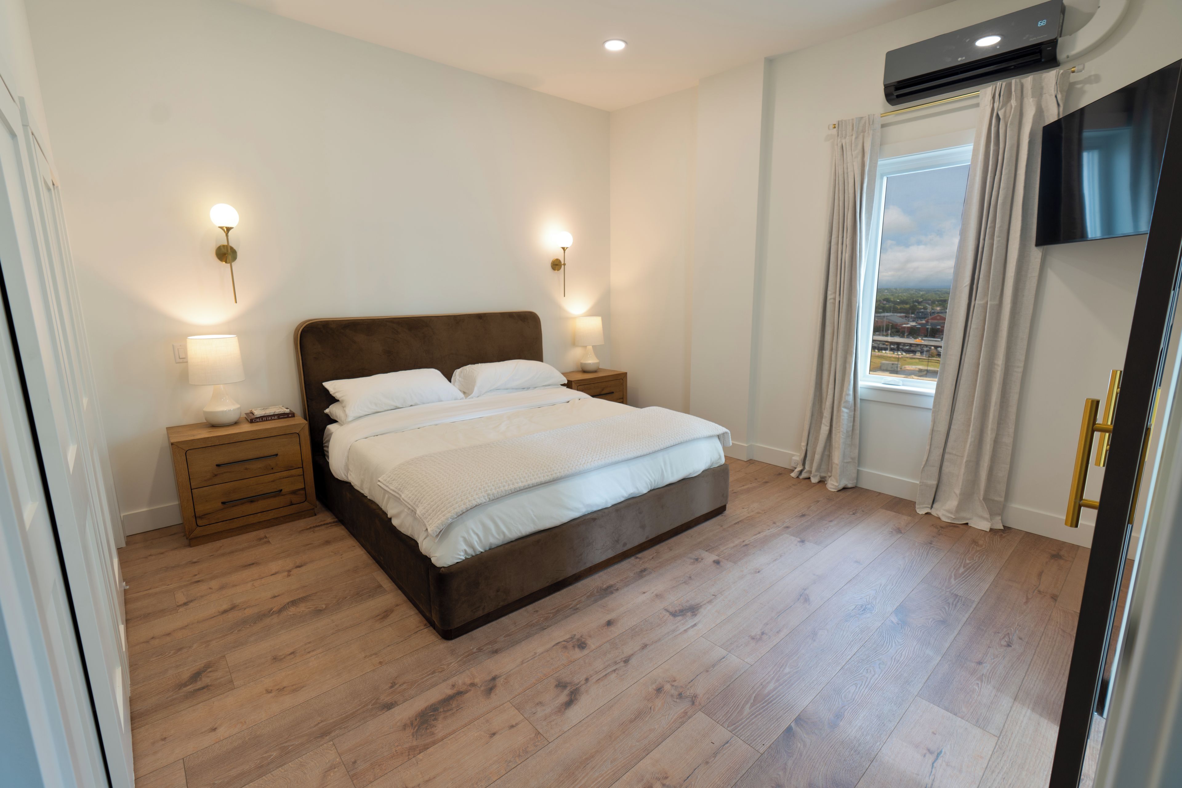 A bedroom with a large bed, bedside lamps, a wooden nightstand, and a view through a window with curtains.