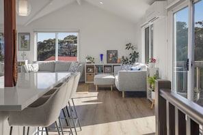 A modern living room with a large window, a dining area with a bar, and a seating area with a sofa and ottoman. - Luxury by the lighthouse (Surf Coast)