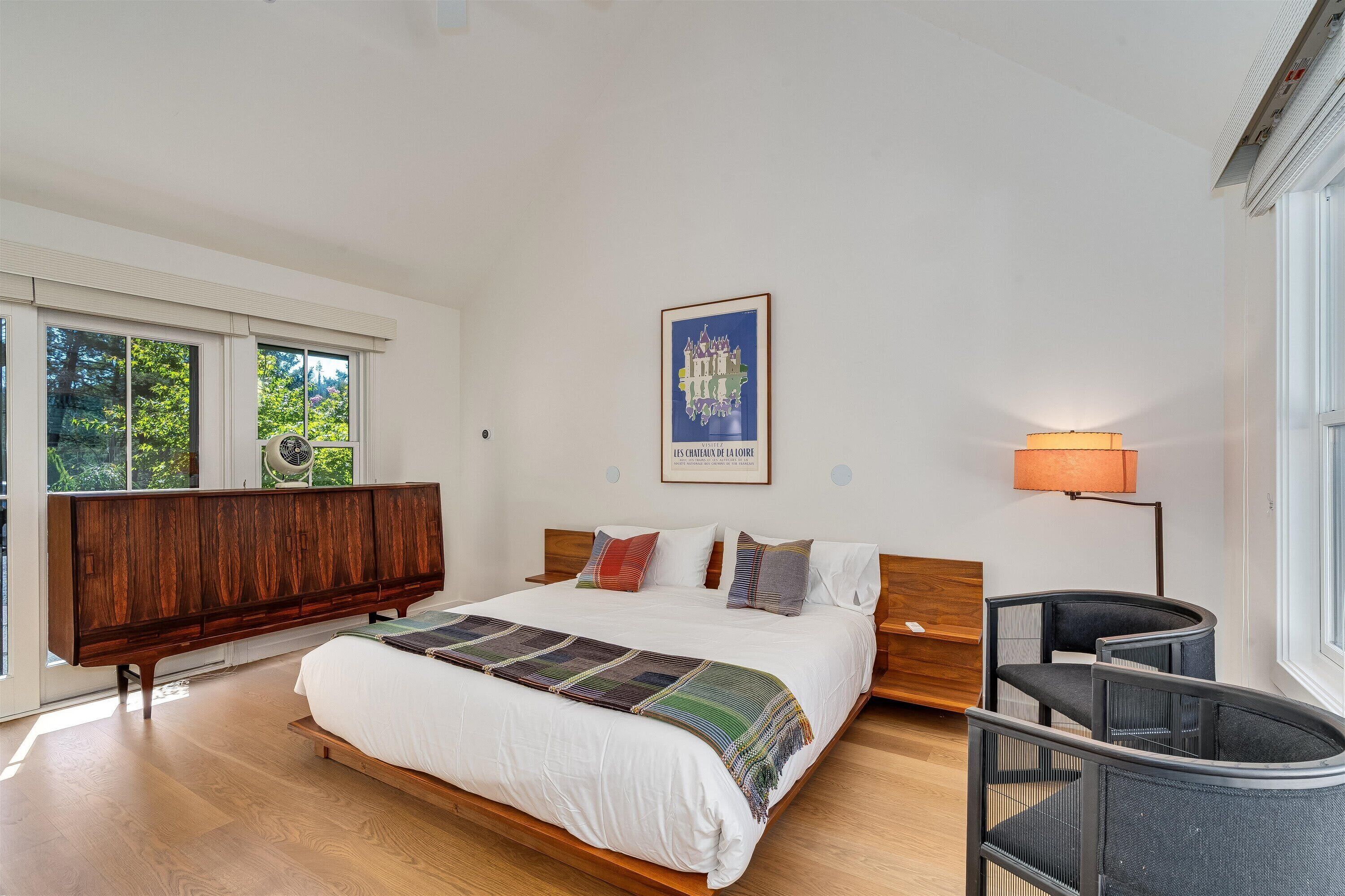 Ein modernes Schlafzimmer mit einem großen Bett, einem Holz-Kopfteil, einem Nachttisch, einem Sessel, einer Stehlampe und einem Fenster mit Blick auf Grünflächen.