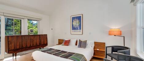 Una camera da letto moderna con un letto grande, una testata in legno, un comodino, una sedia, una lampada da terra e una finestra con vista su piante.