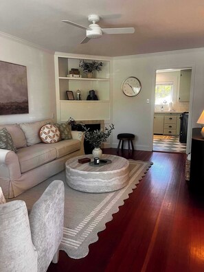 A living room with a sofa, coffee table, and a fireplace. There is a clock on the wall and a ceiling fan. - Plaza St Bungalow -Walk to Plaza, Tasting Included (Healdsburg)