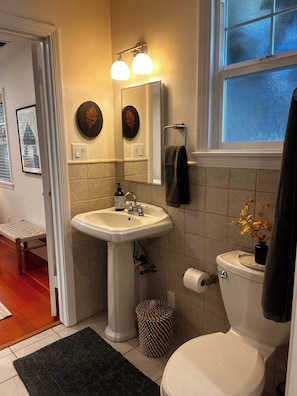 Ein Badezimmer mit einem weißen Waschbecken, einem Spiegel und einer Toilette. Ein Fenster bietet Blick ins Freie.