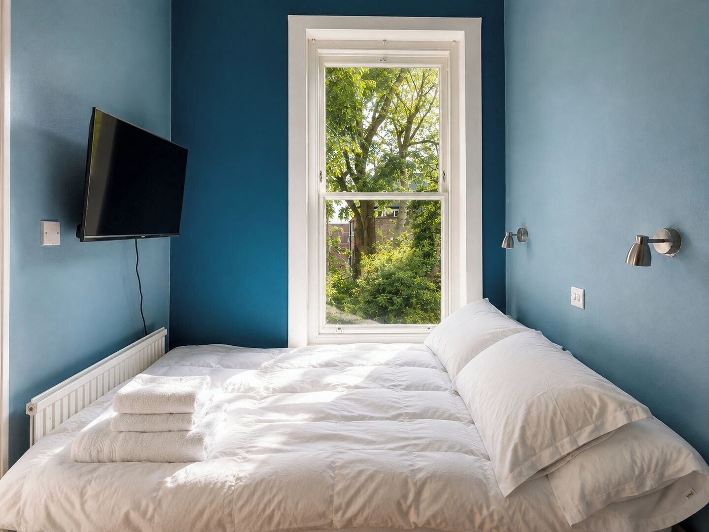 Een slaapkamer met een bed, een aan de muur bevestigde televisie, een raam met uitzicht op groen en een muurlamp.