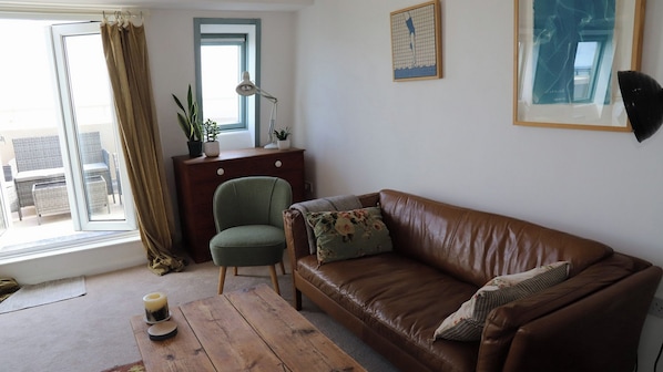 A room with a brown leather sofa, a green armchair, a wooden table with candles, a small wooden cabinet, and a window with curtains.