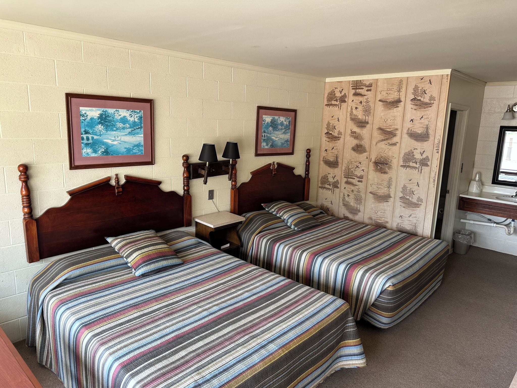 Quarto de hotel com duas camas, mesa de cabeceira, abajur, porta-retrato e parede com papel de parede de tema natural.