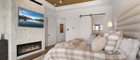 A bedroom with a fireplace, a bed with a plaid bedspread, a ceiling fan, and a large window.