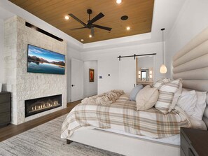 A bedroom with a fireplace, a bed with a plaid bedspread, a ceiling fan, and a large window. - Timpanogos View Estate at Red Ledges (Heber City)