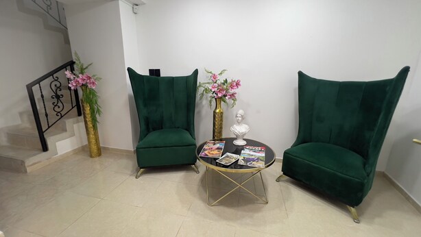 Quarto com duas poltronas verdes, uma mesa redonda pequena com revistas, um vaso dourado com flores cor-de-rosa e uma escada com corrimão preto.