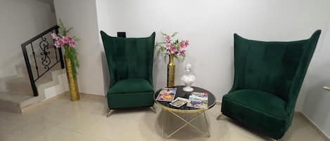 Een kamer met twee groene fauteuils, een klein rond tafeltje met tijdschriften, een gouden vaas met roze bloemen en een trap met een zwarre balustrade.