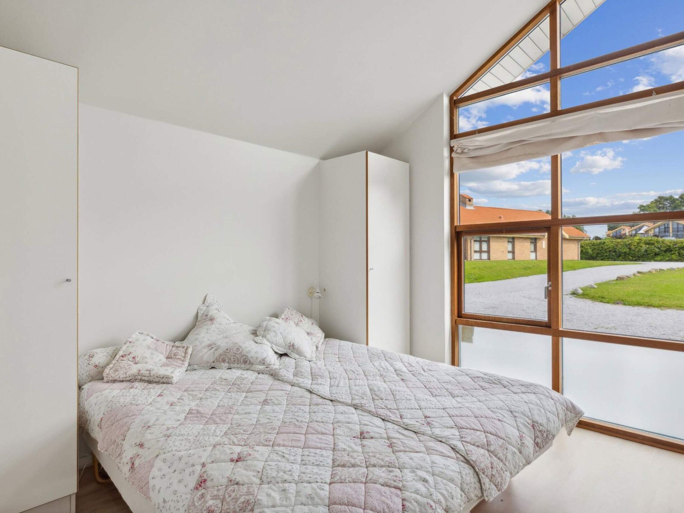 Quarto com uma janela grande, cama com roupa de cama floral e vista para um edifício e área verde lá fora.