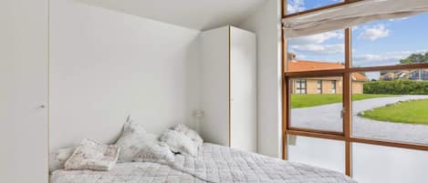 Un dormitorio con un ventanal grande, una cama con ropa de cama floral y vistas a un edificio y vegetación.