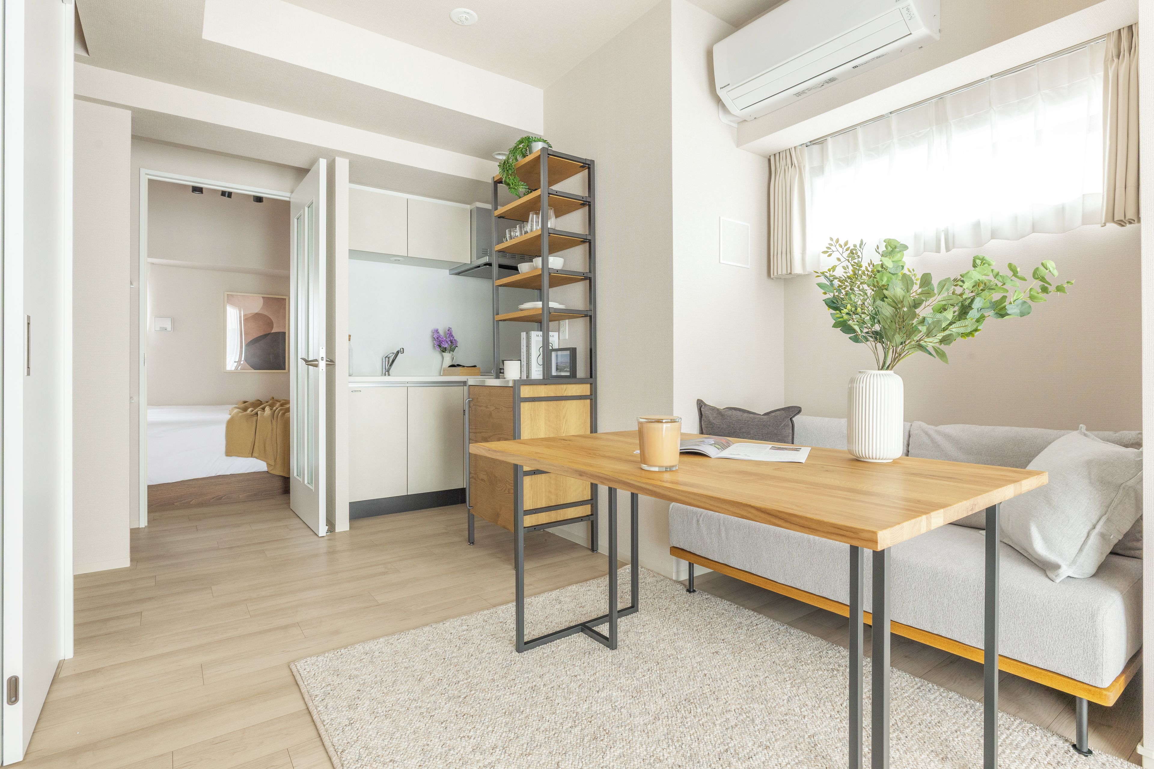 A modern, well-lit living space with a wooden dining table, a sofa, and a kitchen area with open shelving.