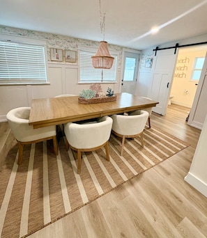 A dining area with a wooden table, four chairs, a woven rug, and a hanging light fixture. - Hibiscus Hideaway - Coastal Retreat by the Beach (Ormond Beach)