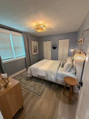 Quarto com cama, abajures, tapete, cômoda de madeira e janela com persianas.