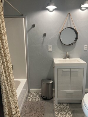 Casa de banho com lavatório branco, espelho, lixeira e chão estampado.