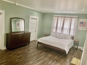 Quarto com cama de madeira, cômoda, espelho, porta e janela com cortinas.