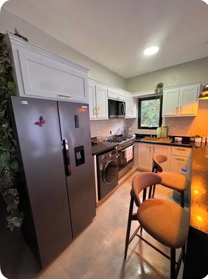 Een compacte keuken met een koelkast, oven, magnetron en een spoelbak. Twee barkrukken en een raam zorgen voor natuurlijk licht.