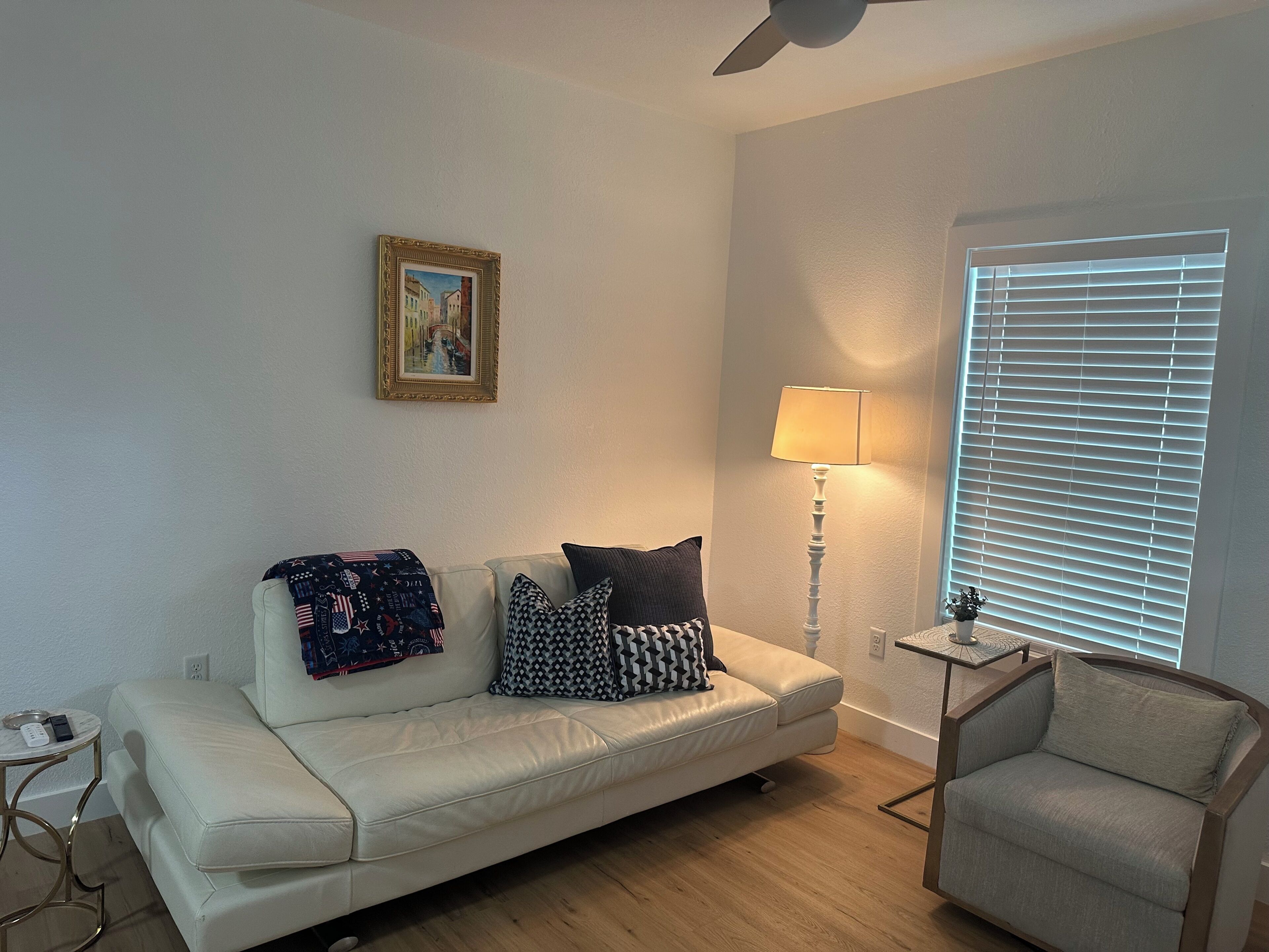 A living room with a white sofa, a small side table, a lamp, a framed picture on the wall, and a window with blinds.