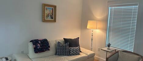 A living room with a white sofa, a small side table, a lamp, a framed picture on the wall, and a window with blinds.