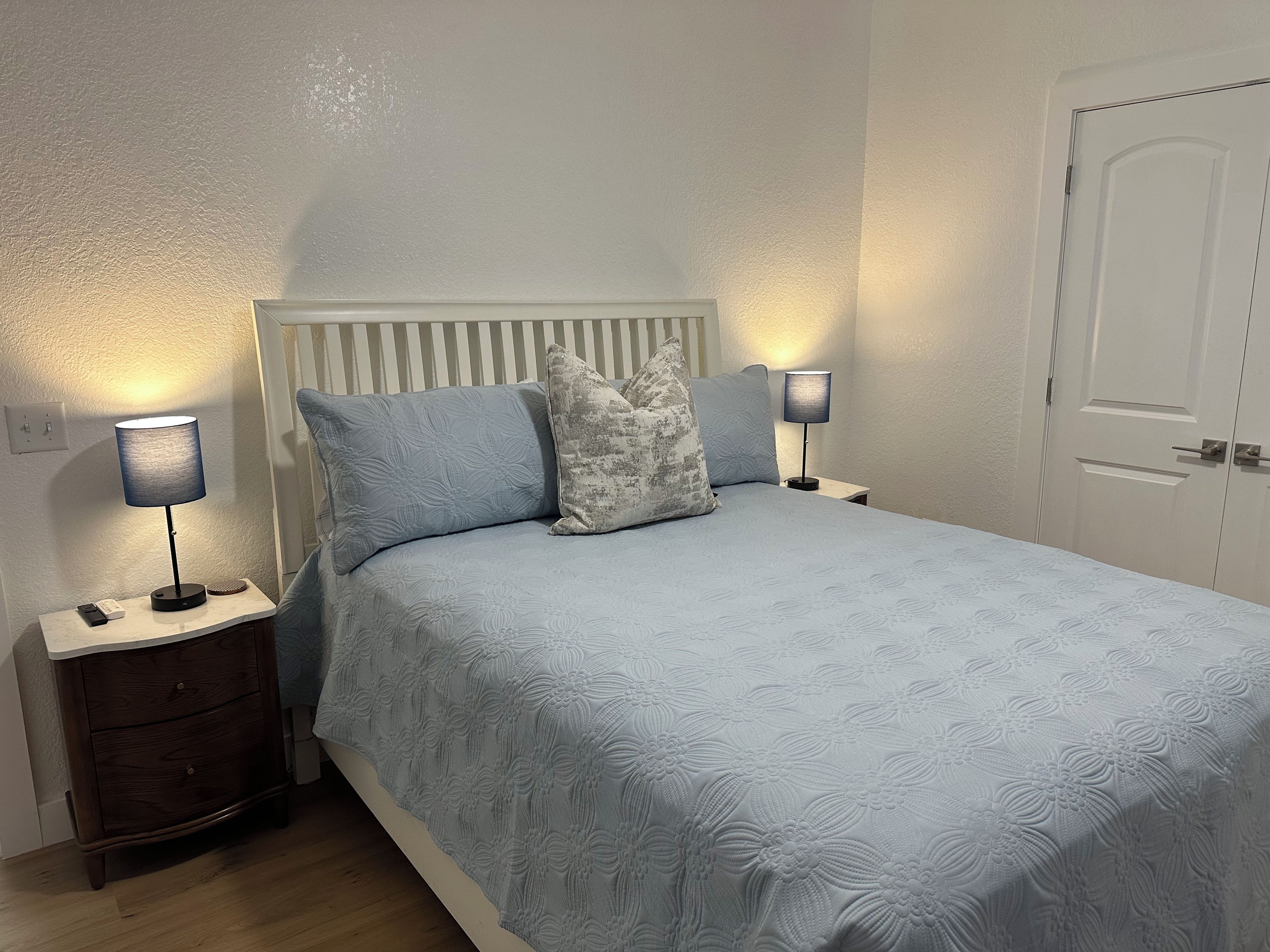 Ein ordentlich bezogenes Bett mit einem blumigen Überwurf, begleitet von zwei Nachttischlampen auf Holz-Nachttischen, und eine weiße Tür im Hintergrund.