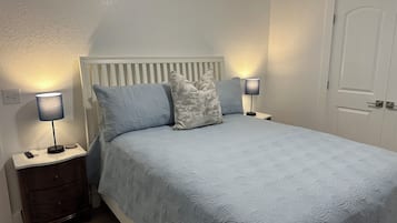 Uma cama bem arrumada com colcha estampada de flores, com duas luminárias laterais sobre mesas de cabeceira de madeira, e uma porta branca ao fundo.