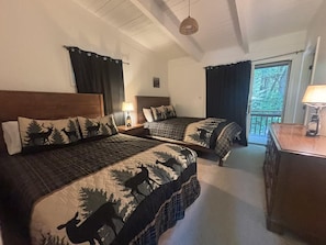 Una habitació amb dos llits, cadascun amb capçalera i un edredó amb motius de bosc. Hi ha una petita taula de nit amb una làmpada entre els llits, i una finestra amb cortines.