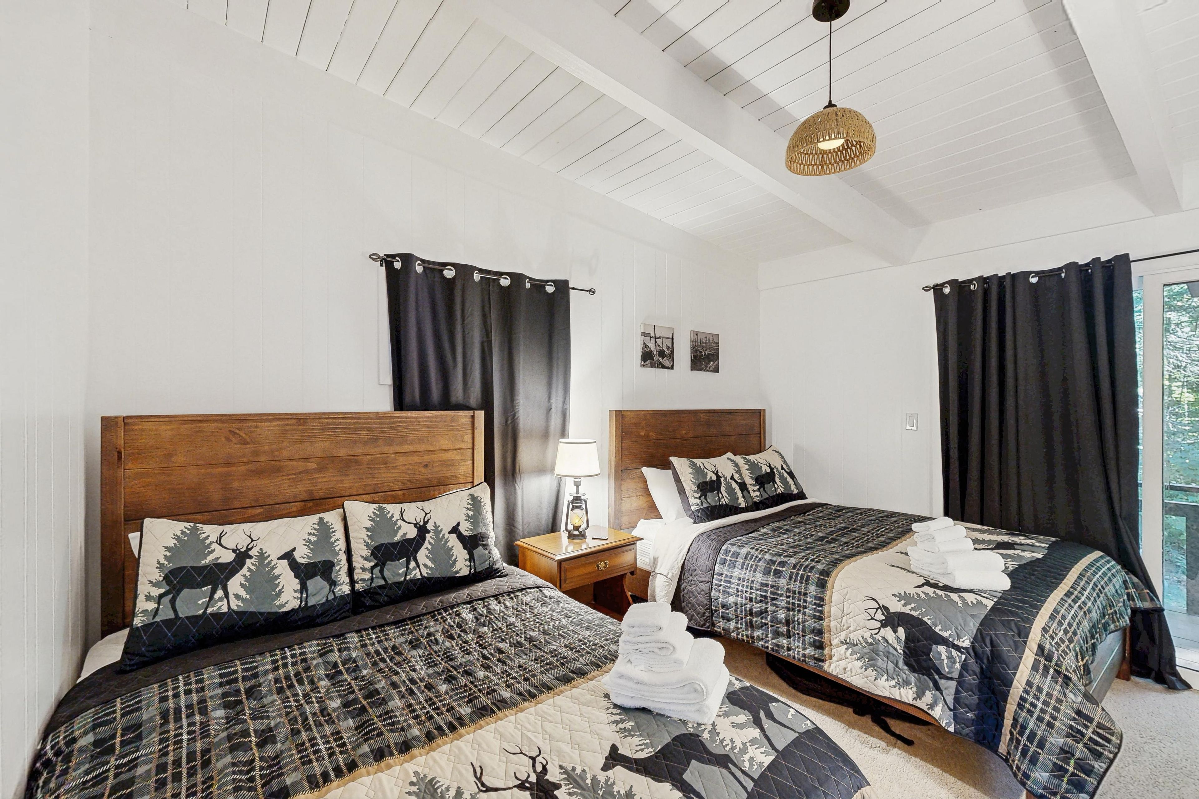 Ein Hotelzimmer mit zwei Betten, jeweils mit Kopfteil und einer gesteppten Bettdecke mit Hirschmuster. Ein Nachttisch mit Lampe, ein Fenster mit Vorhängen und Blick ins Freie.