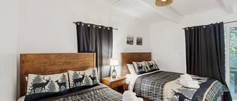 Ein Hotelzimmer mit zwei Betten, jeweils mit Kopfteil und einer gesteppten Bettdecke mit Hirschmuster. Ein Nachttisch mit Lampe, ein Fenster mit Vorhängen und Blick ins Freie.