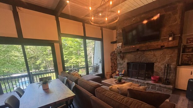 Un salon chaleureux avec une cheminée en pierre, un coin repas avec une table et des chaises, et de grandes fenêtres donnant sur une forêt.