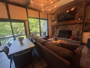 Un salon chaleureux avec une cheminée en pierre, un coin repas avec une table et des chaises, et de grandes fenêtres donnant sur une forêt.