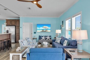 A living room with a blue sofa, a coffee table, and a TV. There is a kitchen area with a refrigerator and cabinets. - Canal Home: Private Pool, Hot Tub, Dock and Pilings (Marathon)