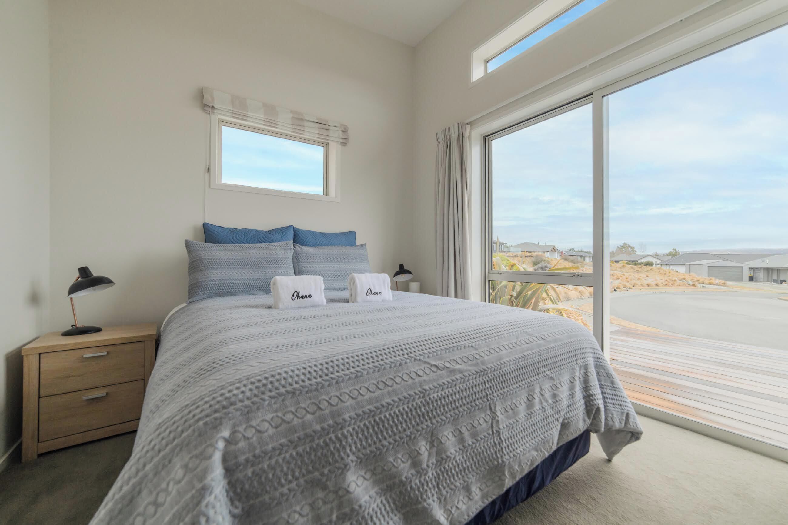 A bedroom with a large bed, two pillows, a nightstand, a lamp, and a window with a view of the beach.
