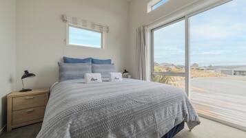 A bedroom with a large bed, two pillows, a nightstand, a lamp, and a window with a view of the beach.