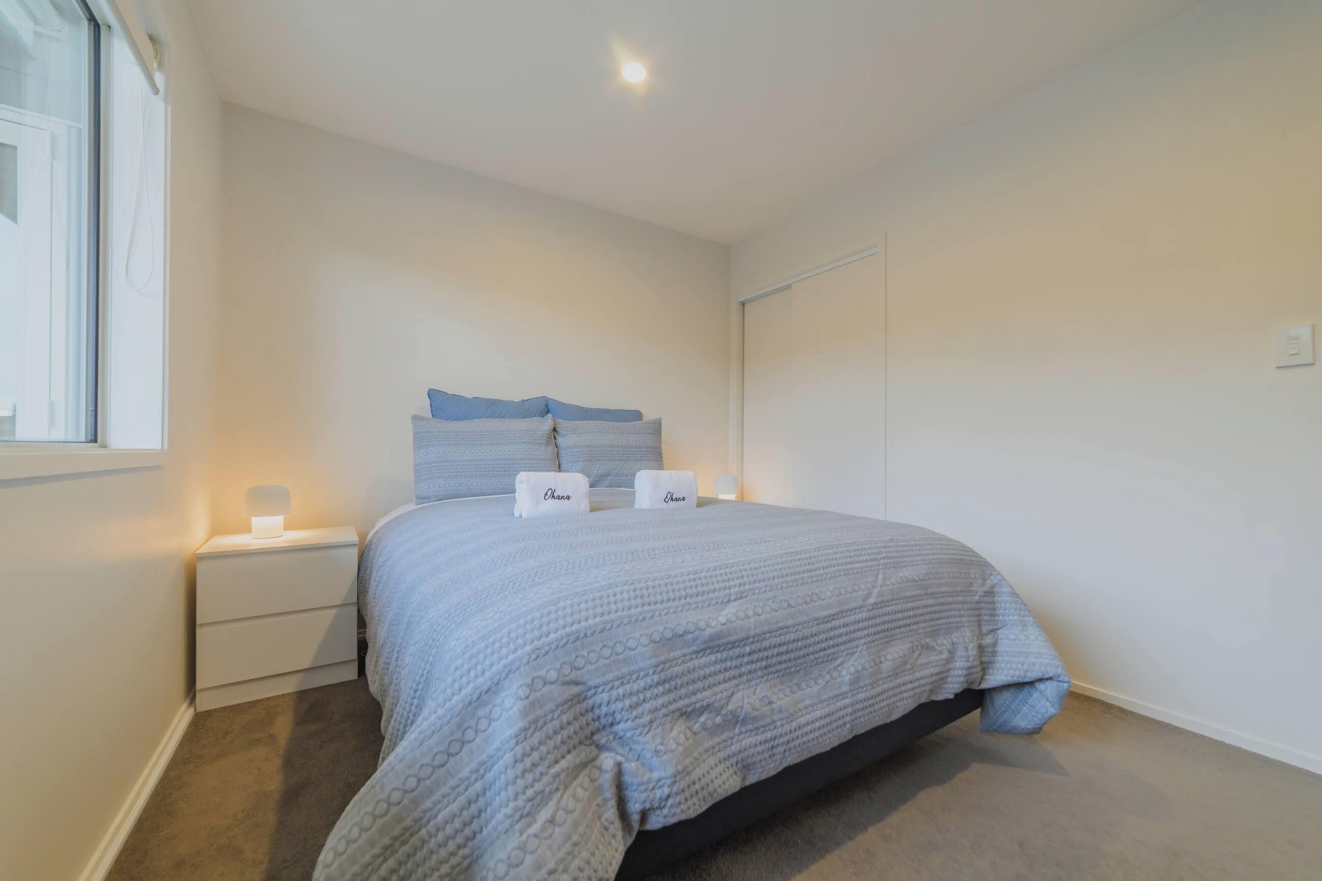 Een slaapkamer met een bed, twee kussens, een nachtkastje met een lamp en een raam.