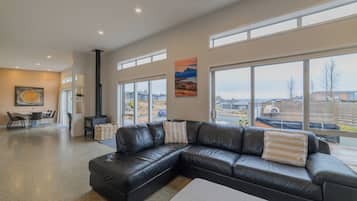 A spacious living room with a black leather sectional sofa, a dining area with a table and chairs, and large windows offering a view of the outside.