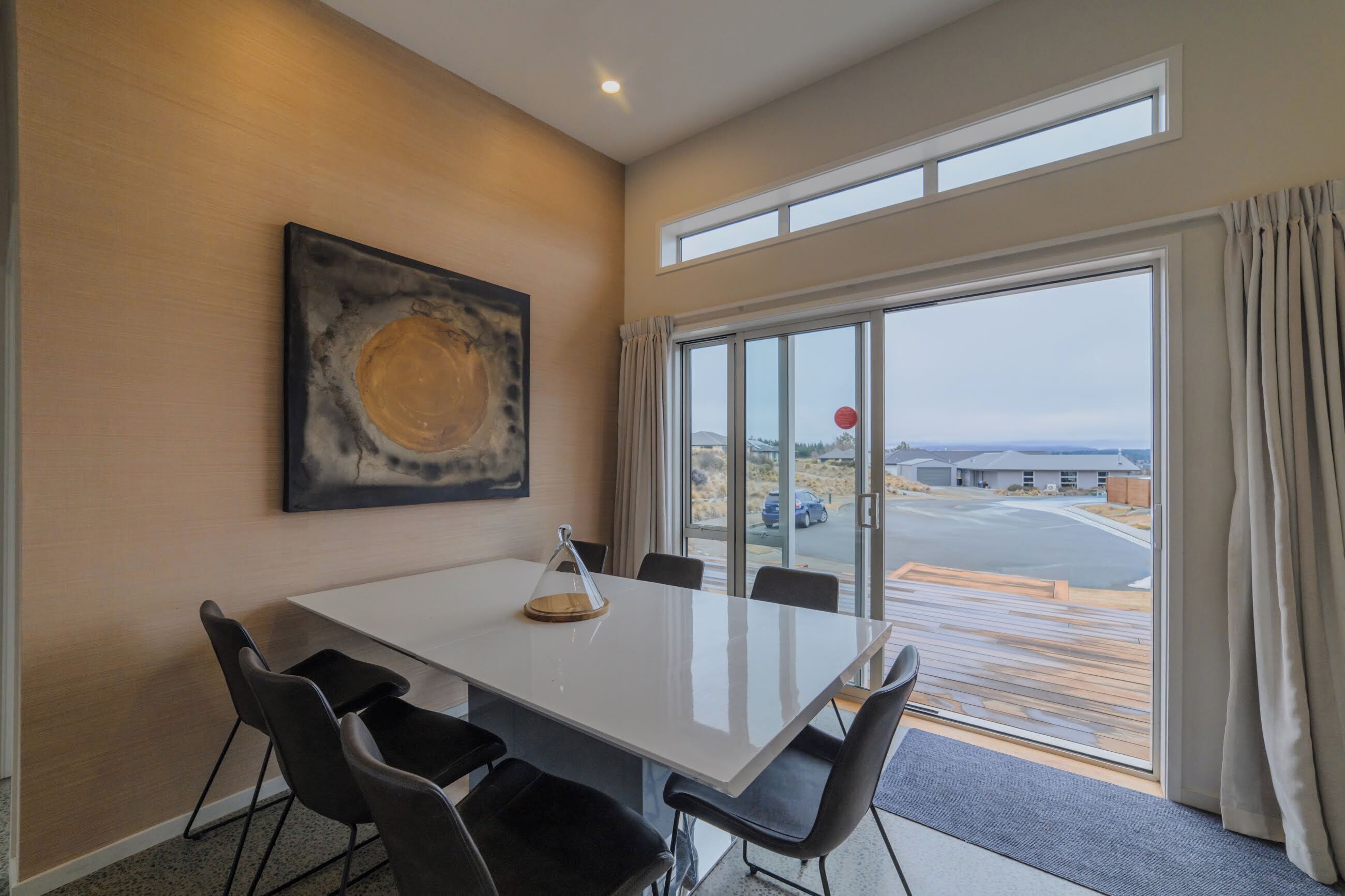 A modern dining area with a white table, black chairs, a glass enclosure, and a large window with curtains.