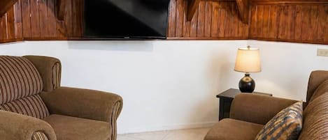 Un salon chaleureux avec un plafond en bois en forme de voûte, un téléviseur à écran plat fixé au mur et deux canapés ornés de coussins à motifs.