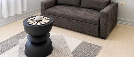 A living room with a grey sofa, a black coffee table with a decorative top, a wall-mounted fan, and a window with white curtains.